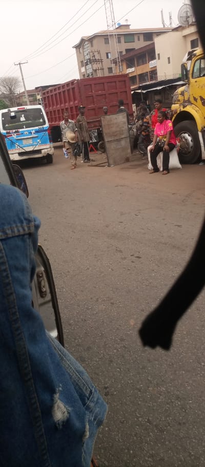 The image depicts a bustling street scene in Ogbunike, Nigeria, observed from inside a vehicle. In the immediate foreground, the left leg of an individual wearing distressed blue denim jeans is partially visible, with a fragment of a white vehicle door frame to its right. The asphalt road surface, marked with minor imperfections, occupies the lower and central portions of the frame.
In the midground, a white and blue commercial minibus, displaying "269" and "NIGERIA" on its rear, faces away from the viewer on the left. To its right, a large red dump truck is parked or stationary. Several individuals are present on the street: a man in a light-colored shirt and trousers walks towards the left, carrying a straw hat; another man in a patterned garment stands near the red truck. Further right, a woman in a bright pink "Arsenal" branded shirt and dark trousers sits on a white sack beside a yellow truck, which is partially visible on the far right. A two-panel metal barrier stands near the center.
The background features several multi-story concrete buildings, some appearing commercial with visible signs, including a partial "Mobil" logo. Rooftops are equipped with satellite dishes and antennas. A prominent tall, open-frame communication tower and multiple utility poles with overhead wires are also visible against an overcast, light grey sky.