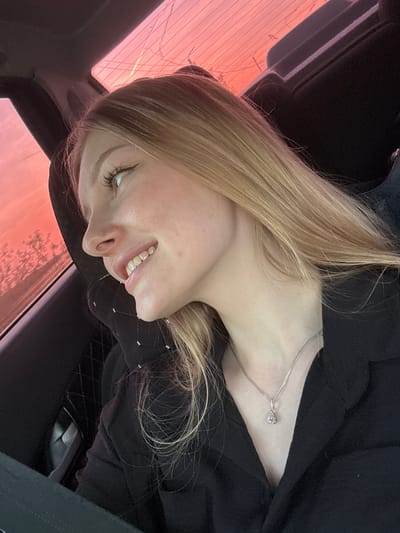 A woman with blonde hair, wearing a black top and a silver necklace with a pendant, is sitting in what appears to be a car. She is smiling and looking off to the side. The background visible through the car window shows a pink and orange sky, possibly during sunset or sunrise. The location appears to be Кондратово, Russia based on the provided instructions, although specific geographic details are absent. The interior of the car is predominantly dark.