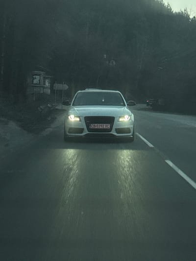 A light-colored Audi sedan, bearing Bulgarian license plate "CB 0392 XC," is centered on a dark asphalt road in Vlado Trichkov, Bulgaria. Its headlights are illuminated, casting two distinct beams onto the road surface. A solid white line marks the right edge of the visible lane. To the immediate left of the road, a dilapidated, light-colored structure with dark window openings is partially visible through dense, dark trees, adjacent to a multi-arrow directional signpost. The background on both sides of the road consists of heavily wooded, dark slopes. In the distant background on the right, another vehicle with faint lighting is discernible. The scene is depicted under low-light conditions, consistent with dusk or dawn. No individuals are present.