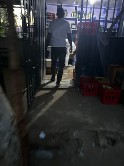 In Mayo Dasa, Nigeria, a person wearing a white t-shirt and dark trousers walks away from the camera, entering a store. The store shelves are stocked with bottles, and red plastic crates are visible outside the entrance. The lighting suggests it is either dusk or nighttime, casting long shadows on the ground.