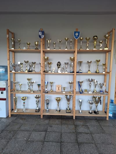 The image depicts an indoor scene in Ezerovo, Bulgaria, showcasing a multi-tiered wooden shelving unit displaying approximately 60-70 sports trophies. The unit, constructed from light-colored wood, comprises four vertical support sections and five horizontal shelves positioned against a plain, light-grey wall.
The trophies are diverse in design, predominantly metallic with gold, silver, and bronze-toned finishes. They include classic cup shapes, figurines (such as a stylized footballer), and spherical tops resembling a football. Many trophies rest on dark bases, some with legible inscriptions.
Specific elements on the shelves include:
* On the upper left shelf, a blue and white shield-shaped pennant is partially visible, bearing the Cyrillic text "СПОРТНА АКАДЕМИЯ БОРИСЛАВ" (Sports Academy Borislav).
* On the upper right shelf, a blue and white triangular pennant with a crest is displayed, reading "АКАДЕМИЯ НАЦИ" (Academy Naci).
* On a lower shelf, a small brown rectangular plaque with "LONGOMATCH" and "II МЯСТО" (2nd Place) is present.
* One trophy on a middle shelf features a blue ribbon attached.
To the far left, a white water dispenser with a red spigot is visible, topped by a large, clear blue 5-gallon water bottle labeled "AQUA." To the far right, a similar blue 5-gallon water bottle rests on the floor.
The floor is covered with dark grey, speckled concrete or terrazzo tiles, showing visible grout lines and signs of wear. The lighting is consistent with indoor ambient conditions. No individuals are present in the scene.