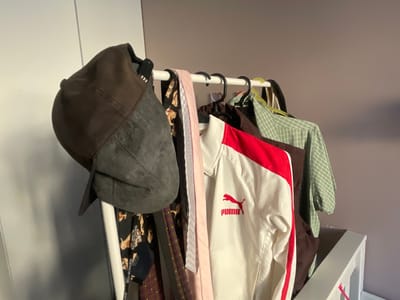 A rack of clothing is displayed against a pale purple wall. On the left, a dark grey flat cap is hung, its brim angled downwards. Next to it, several ties are visible, one with a pink and white checkered pattern and another with a cartoon pattern featuring characters that appear to be from Looney Tunes, with text that reads "BABYMILKSTORE". A white Puma track jacket with red stripes adorns the rack, followed by a dark brown garment and a green checkered shirt. The scene suggests a domestic setting, possibly a bedroom or closet, and the lighting indicates daytime indoors. No weather or specific time of day cues are evident.