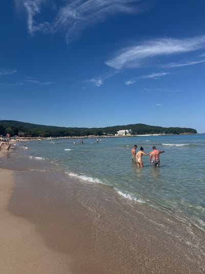 A wide shot captures a sun-drenched beach in Primorsko, Bulgaria, with crystal-clear turquoise waters gently lapping the shore. The sand, a light tan color, stretches from the foreground towards a line of lush green trees and hills that form the backdrop. A few distant buildings are visible on the hilly landscape.
In the foreground, three individuals stand in the shallow water. A man on the left, wearing patterned swim trunks, faces away from the camera. Next to him, a woman in a patterned bikini faces left, her left arm resting on the man's right arm. To the right of the woman is a man in patterned swim trunks, his back to the camera, with his right arm extended forward as if pointing at something in the distance.
Further out in the water, numerous other people are scattered across the scene, some wading, others swimming or engaging in water activities. A few surfboards are visible. The sky above is a vibrant blue, dotted with wispy white clouds, indicative of a clear summer day. The overall impression is one of a relaxed and popular seaside resort.