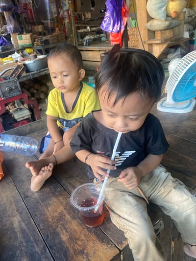This media file captures a candid moment of two young boys, likely brothers, in an indoor setting with a rustic wooden floor. The older boy, wearing a black t-shirt, is lying down and drinking from a clear plastic cup with a white straw. His eyes are closed, suggesting relaxation or sleepiness. The younger boy, in a yellow t-shirt, is sitting beside him and looking towards the camera with a neutral expression.

The scene appears to be a home or a simple living space, indicated by the presence of various objects in the background. To the left, there's a blue and white electric fan, along with some stuffed toys and what looks like wooden shelving units. Scattered items, including plastic bags and what could be produce, suggest a bustling, perhaps domestic, environment. A partially visible water bottle and another cup with dark liquid are also present on the floor.

The lighting suggests it could be daytime, though the exact time of day and weather are not discernible. The overall atmosphere is informal and intimate, highlighting a moment of quiet rest or refreshment for the children. The wooden floor and the assortment of items hint at a lived-in space, possibly in a rural or less modernized area. No specific text is legible in the image.