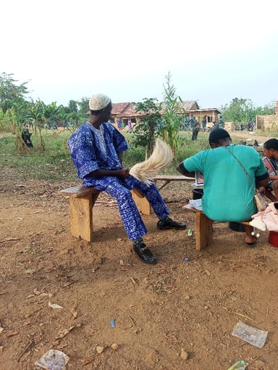 This outdoor photograph depicts a lively scene in Aramoko-Ekiti, Nigeria, under a bright daytime sky, possibly in the late afternoon.

In the left foreground, a man sits on a simple wooden bench, dressed in a vibrant blue and white patterned traditional Nigerian outfit, which includes a flowing tunic and matching trousers. He wears a white, textured skullcap and shiny black loafers. The man is positioned with his back partially to the camera, looking towards the background. He holds a light-colored, possibly cream or white, whisk or fly swatter, often used as a cultural accessory or for practical comfort.

To the right, another person is seen from behind, seated on a similar wooden bench. This individual wears a teal or green t-shirt and dark trousers, with a patterned shoulder bag slung across their back. A third person is partially visible next to them on the far right. These individuals appear engaged with items on the bench or table in front of them.

The ground is unpaved, consisting of dry dirt with scattered debris, including plastic wrappers and crumpled paper. A faded Nigerian Naira banknote is faintly visible on the ground in the bottom right corner, though its specific denomination is illegible.

The mid-ground and background feature several rustic buildings with corrugated iron roofs, nestled amidst green foliage and tall, slender crops, possibly corn or sorghum. Numerous people, some appearing to be children or students in blue patterned uniforms similar to the main subject's attire, are visible near these structures. Their presence suggests a community gathering, school activity, or a bustling local scene. The overall impression is one of communal activity in a rural or semi-urban setting.