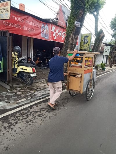 A man pushes a "KUE PUTU KLEPON" food cart along a street. The cart is wooden with two wheels and a small cabinet. He wears a blue shirt, light-colored pants, and flip-flops. In the background, a motorcycle repair shop displays a red banner that reads "motor matikmu Spesialis dingin?" A scooter sits parked in front of the shop. Further down the street, trees line the sidewalk, and signs are visible on nearby buildings.