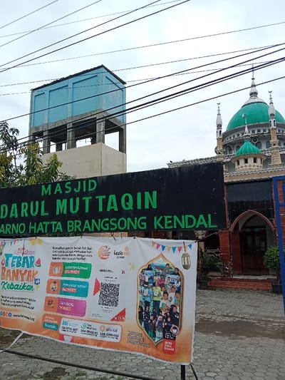 The image shows a mosque in the city of Kecamatan Brangsong, Indonesia, at the coordinates -6.948 latitude and 110.23 longitude. The mosque has a green dome and minarets, and is located in a residential area. The mosque is surrounded by a brick wall and a gate with a black sign that says "MASJID DARUL MUTTAQIN ARNO HATTA BRANGSONG KENDAL". There is a water tower to the left of the mosque, and a large banner in front of the mosque, with a QR code on it. The banner also says "Ramadhan penuh berkah, Yay Mu akan senantiasa hadir untuk berkontribusi, bersama Lazismu Kendal, dengan cara transfer atau iuran derma..." in Indonesian. The background is a cloudy sky. There are no people visible in the image.