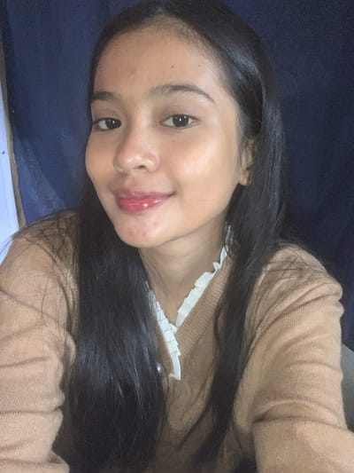 The image is a close-up selfie of a young woman with dark hair, facing the camera. She is wearing a tan cable-knit sweater over a white ruffled-collar top. Her lips are glossy and appear to have red lipstick. The background is a dark blue fabric. The image does not provide any information about the location in Sungai Meriam, Indonesia.