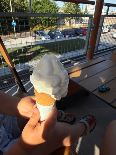 The image captures a close-up, elevated view of a person, likely a child, enjoying a white soft-serve ice cream cone outdoors in Burjassot, Spain. The focus is on the child's hands, which cradle the waffle cone and its patterned paper sleeve, with strong sunlight highlighting the skin and casting deep shadows. The ice cream itself is a smooth, white swirl.

The child is seated at a wooden outdoor table or bench, where a small blue and green toy car is visible. The bright sunlight creates clear contrasts and long shadows across the table surface. The person's lower legs and feet, clad in casual shoes (possibly sandals or sneakers) with distinctive orange and blue laces, are also visible.

Beyond the immediate foreground, a metal mesh fence or railing separates the scene from an outdoor street or parking area below. Through the fence, several cars of various colors (red, blue, silver, white) are visible on a paved surface. Lush green trees and foliage line the street, and distant buildings can be seen. A blue circular road sign with a white arrow pointing upwards is visible through the fence, indicating a "straight ahead" direction.

The presence of bright sunshine and strong shadows suggests a clear, sunny day, likely during mid-morning or late afternoon, creating a pleasant atmosphere for an outdoor treat. The setting appears to be an elevated outdoor space, such as a balcony or terrace, overlooking the street below.