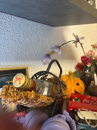 This indoor scene, located in the city of Kuklen, Bulgaria, presents a collection of decorative and functional objects arranged on a surface, likely a shelf or cabinet, against a textured wall. The wall, appearing light in color, features a coarse, stucco-like finish. Above the wall, a flat, gray surface suggests the underside of another shelf or an overhead structure.
In the foreground, a clear glass bottle with a gold-colored screw cap and a red band is prominently displayed. The cap is embossed with a red crown-like symbol flanked by two stars. The bottle rests within a woven, light-brown basket made of natural fibers, which is itself contained within a dark, possibly wrought-iron, decorative stand with intricate scrollwork. Beneath the basket, a small section of a gold-framed picture, possibly depicting a tropical landscape, is visible.
To the right of the bottle arrangement, an orange jack-o'-lantern style pumpkin decoration is partially visible, showing carved features for eyes and a mouth. Further right, a dark, ribbed vase holds a small bouquet of artificial flowers, predominantly pink and red, with some white accents. Behind and above these elements, a slender green stem with light purple artificial flowers extends upwards and across the scene.
Below the main display, several objects are clustered. Two red boxes, clearly branded "Coca-Cola" with the classic logo and an image of a Christmas ornament, are stacked. To their left, a small, dark blue toy car or van is visible, draped with a light-colored beaded necklace. In the immediate foreground, two padded, purple objects, possibly parts of headphones or similar accessories, are present, along with a small pink box labeled "parfum bg". The overall lighting is soft and consistent, indicating an indoor environment.