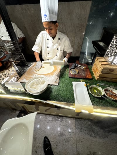 An eye-level indoor shot captures a male chef in a white uniform and toque, diligently preparing dough with a rolling pin on a wooden board. He wears white gloves while meticulously working on a circular piece of dough. The text "GALIA" in blue, accompanied by a wave-like logo, is visible on his chef's hat.

The chef stands before a counter styled with artificial green grass, upon which various ingredients and tools are arranged. To the right of his workspace, a large bowl filled with white flour is present. Further to the right, two white hexagonal bowls contain red chili flakes and a vibrant green sauce, possibly pesto, each with a spoon. A pizza cutter and another spoon rest on a smaller wooden cutting board near the prepared ingredients. A long, empty white serving dish sits along the right edge of the counter, illuminated by a strip of LED lights running beneath it.

The setting appears to be a live cooking station or buffet within a modern restaurant or hotel, likely in New Town, India, given the provided context. The background features dark, patterned walls or decorative panels, and a reflective surface directly below the chef, reflecting parts of his attire. Artificial lighting dominates the scene, suggesting an evening or night-time event. The combination of culinary activity and decorative elements like wooden crates and artificial grass creates an engaging and functional display.