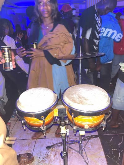 A low-angle, vertical shot captures a vibrant scene in what is identified as the city of Wumat, Nigeria. In the foreground, a pair of bongos rests on a stand, their surfaces appearing worn. A cymbal is suspended above them. To the left, a person's hand holds a can, partially obscuring a woman in a brown and blue outfit who is looking towards the right. Behind her, a man in a black shirt with a white skeleton-like print and a gold chain is visible. To his right, another person wears a bright blue jacket with the word "supreme" emblazoned on it. The environment appears to be indoors, possibly a club or event space, with blue lighting casting a cool glow on the scene. The floor is tiled with a combination of light and dark patterns. Other individuals are present in the background, though their details are less distinct.