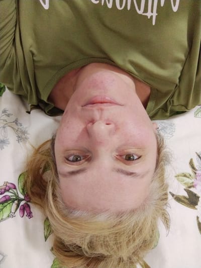 The image shows an upside-down, close-up selfie of a woman lying on a floral-patterned surface. She has blonde hair, light-colored eyes, and is wearing a green t-shirt. The background appears to be a bed or sofa with a light-colored fabric featuring a botanical print of flowers and leaves. The lighting suggests an indoor setting, possibly a bedroom. There are no discernible landmarks or clues to indicate the specific location within Al Mas'oudi, United Arab Emirates. The image focuses solely on the woman's face and upper torso.