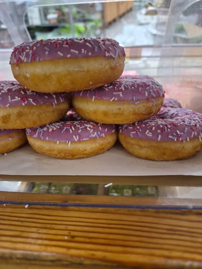 A stack of donuts with purple icing and sprinkles is displayed on a wooden surface. The donuts are arranged in a pyramid shape, with three donuts on the bottom layer and two on the second layer, and one on top. The donuts are golden brown on the sides and have a ring shape. The icing is a light purple color and is topped with small, multicolored sprinkles. The background is blurred and appears to be an indoor setting, possibly a bakery or market. The image is a close-up shot, focusing on the donuts. The donuts are likely fresh and ready for consumption. The scene appears to be from a display case, suggesting a commercial setting. The overall impression is one of delicious and visually appealing pastries.