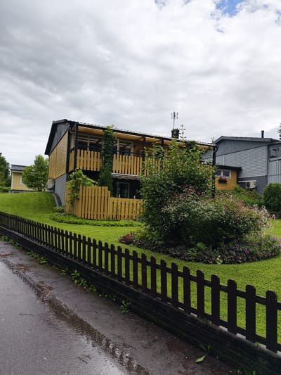 The image depicts a residential area in Ljungsbro, Sweden, under an overcast sky. In the foreground, a dark wooden picket fence borders a wet, asphalt road that reflects the sky and surrounding greenery. Beyond the fence, a neatly mown green lawn slopes upwards towards a multi-level wooden house painted primarily in yellow and black. This house features a covered balcony with yellow wooden railings and a dark roof. Lush green bushes and flowering plants surround the base of the house and spill onto the lawn. To the right of the yellow house, a grey, two-story building with horizontal siding is visible, featuring a small yellow annex with a window box. An outdoor air conditioning unit is mounted on the wall of the grey building. The scene suggests a typical suburban or rural residential setting, possibly after a rain shower given the wet road. No people are visible in the image.