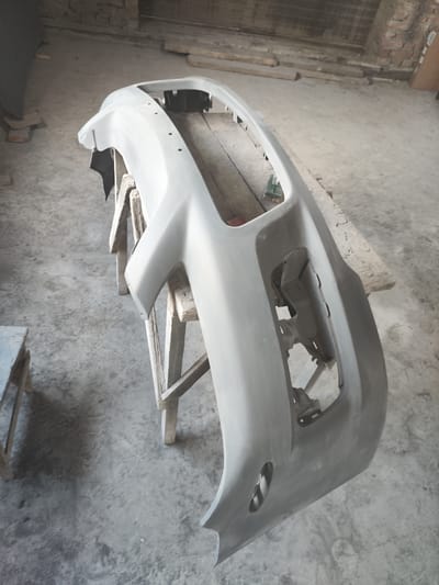 A car bumper, primed and unpainted, is elevated on a wooden sawhorse. The bumper is angled towards the right, with its front facing the viewer. It has cutouts for lights and vents. The environment appears to be an indoor workshop or garage, with a concrete floor. Wooden planks and brickwork are visible in the background. There are no people present in the image. The scene is lit by ambient light, creating shadows on the floor.
