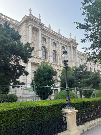 The image shows the facade of a large, ornate, white building in Vienna, Austria, located at the coordinates 48.203, 16.362. The building is made of stone and has many windows and columns. There are several statues on the top of the building. The building is surrounded by trees and bushes. In front of the building, there is a metal fence and a black lamppost with a single light.  The light is off and the lamppost is made of ornate metal. The ground is paved with stone. The scene appears to be taken during the day. There are no people in the image.