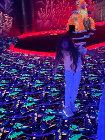 The image depicts an indoor, blacklight-illuminated environment, likely a glow-in-the-dark mini-golf course or entertainment venue, located in Columbus, United States of America. A child, viewed from behind, stands in the middle ground, facing into the brightly glowing scene. She wears a light-colored top, light blue pants featuring heart-shaped cutouts or designs near the ankles, and dark shoes with luminescent soles. Her long, dark hair flows down her back.
In the foreground, to the left of the child, is a large, textured object resembling an abstract coral reef or rock formation. This structure is predominantly orange and yellow with accents of blue and green, including small blue coral-like growths and elongated green shapes. It has dark recesses and a somewhat organic appearance.
The entire space is bathed in ultraviolet light, making fluorescent materials glow intensely. The floor and walls are covered in an intricate, repeating pattern of vibrant blue, green, yellow, and orange squiggles and abstract shapes against a dark background. Bright red fluorescent strips line the edges of paths or architectural elements near the child and the coral structure, providing stark color contrast. A glowing blue element is also visible at the top of the frame, possibly part of the ceiling or an overhead decoration. The overall atmosphere is dark yet colorful and playful, characteristic of blacklight attractions. No discernible text is visible within the image.