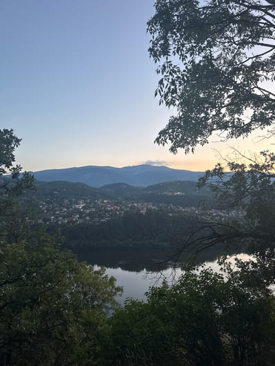 The image captures a panoramic view of Pancharevo, Bulgaria, at dusk. In the foreground, dense green foliage of trees frames the scene, with branches and leaves silhouetted against the sky and distant landscape. A still body of water, likely Pancharevo Lake, occupies the lower portion of the frame, its surface reflecting the muted light of the setting sun and the sky.
Beyond the lake, a town is nestled amidst rolling, forested hills. The houses, with their reddish-tiled roofs and scattered lights, indicate a populated area. The hills are covered in a thick carpet of trees, creating a natural, verdant backdrop.
In the distance, a range of majestic mountains stretches across the horizon. The mountains are bathed in the soft, warm hues of twilight, with subtle variations in shade and texture suggesting their rugged terrain. The sky above is a gradient of clear blue near the zenith, transitioning to pale yellow and orange near the horizon, indicating the last light of the day. The overall atmosphere is serene and tranquil, characteristic of a peaceful evening in a scenic natural setting.
