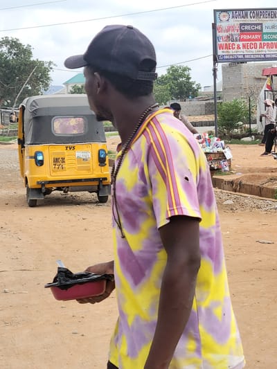 The image shows a man wearing a black cap and a yellow and purple tie-dye shirt, standing next to a yellow tricycle (TVS, registration GGE-444OR, Lagos). He is holding a small red object with black material protruding from it. In the background, there's a sign for "AGHAM COMPREHENSIVE," advertising Creche, Nursery, Primary, and Secondary levels, as well as WAEC & NECO approvals, with the motto "Learning Today, Leading Tomorrow," and phone numbers. The location is identified by the sign as being in Rafinsanyi New Layout, Kwamba, Nigeria. The ground is dirt or sand.
