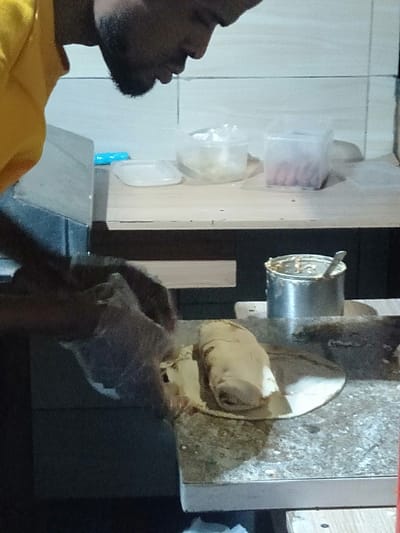 A man wearing a yellow shirt is working on a flatbread on a counter. He is spreading something from a metal can onto the bread. Behind him are various containers on a shelf. The location is Epe, Nigeria.