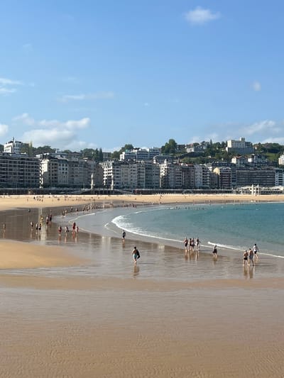 This image captures a wide view of La Concha beach in San Sebastián, Spain, on a sunny day. The foreground shows a wet, sandy beach with ripples from receding water, reflecting the light. Several individuals are scattered across the beach, some walking towards the water, others moving away. In the mid-ground, a gentle wave breaks along the shoreline, with more people visible wading in the shallows and swimming further out. The background features a crescent-shaped bay lined with a promenade and a dense collection of multi-story buildings, typical of the city's architecture. The buildings are a mix of cream, white, and beige, with many windows visible. Behind the buildings, a green, hilly landscape rises, dotted with more structures, and the sky above is a clear blue with a few wispy clouds. The overall scene conveys a sense of a bustling summer day at a popular urban beach.