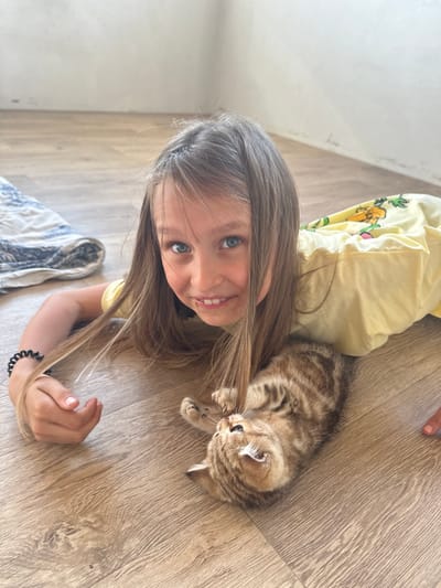 An over-the-shoulder medium shot shows a young girl with long, light brown hair and blue eyes, lying on a wooden floor and looking at the camera with a wide smile. She is wearing a yellow t-shirt with a pineapple print. A small, tabby kitten is lying next to her, on its back, with its paws in the air. The kitten is looking up at the girl, who is reaching out to pet it. The background is a plain white wall. The scene takes place indoors, on a wooden floor, likely in a home environment. It is not possible to determine the specific location in the city of Mokino, Russia from the image alone.