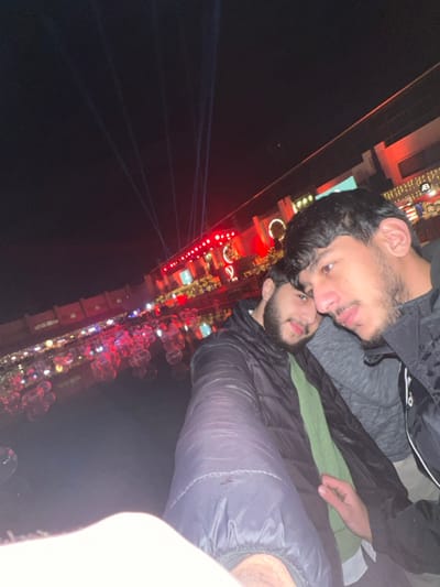 Two young men pose for a selfie at night, with a brightly lit structure in the background. The men are close together, one wearing a dark jacket and the other a grey shirt with a black jacket partially visible. The background includes lights and what appear to be decorative bubbles, along with beams of light shining into the night sky. The image is taken at an angle, with the background appearing to stretch upwards into the dark sky.