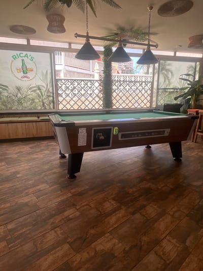 The image shows a pool table in a dimly lit indoor establishment. The establishment appears to be a bar or restaurant, with tropical-themed decor, including plants and woven light fixtures. A sign on the window behind the pool table reads "RICA'S Cocktailbar & Restaurant." The floor is covered in dark, wood-like tiles. There are no people visible in the image. The scene suggests a recreational area within a commercial establishment. The location is identified as Benidorm, Spain, based on the context of the establishment's name and typical tourist offerings in that city.