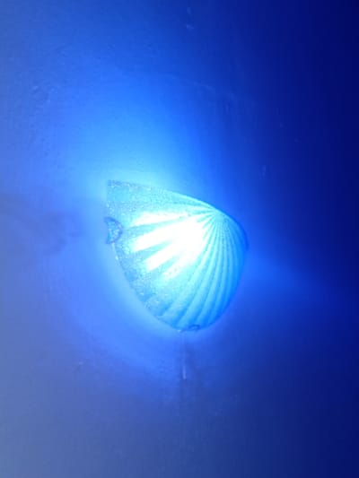 The image shows a close-up of a blue LED light, possibly a safety light or indicator, emitting a bright glow. The light has a ridged or segmented surface, giving it a fan-like appearance. The light is surrounded by a soft blue haze, suggesting it is either underwater, in a dimly lit environment, or the light itself is creating the surrounding illumination. There are small, irregular specks on the surface of the light, which could be water droplets or dust. A subtle, curved shadow or reflection is visible beneath the light, adding depth to the scene. The overall impression is one of a small, functional object within a diffuse, luminous blue environment. There is no visible text in the image. The location context of Pantinapu, Nigeria does not provide any specific visual cues within the image itself.