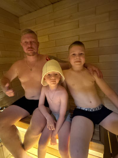 The image depicts three individuals, an adult male and two children, seated on wooden benches in a sauna or banya, characterized by horizontal wooden slat walls and warm, dim lighting. The scene is located in Мишкино, Russia.
The adult male, positioned on the left, is light-skinned with very short blonde hair, a mustache, and a chin beard. He wears a silver-colored chain with a small cross-like pendant and black swim trunks. A tattoo is visible on his upper left arm. His left hand displays a thumbs-up gesture, while his right arm is placed around the shoulder of the older child.
The younger child, situated in the center, is light-skinned and wears a light-yellow felt sauna hat, obscuring his hair. He also wears a silver-colored chain with a small cross-like pendant and light-colored underwear.
The older child, on the right, is light-skinned with short, dark hair. He wears black boxer briefs featuring a visible "Calvin Klein" brand waistband. His left arm extends behind the younger child.
All three individuals are oriented towards the camera.