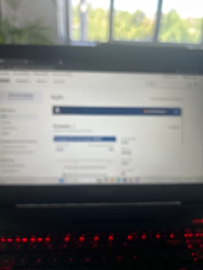 The image shows a close-up view of a laptop screen displaying a financial or accounting interface. The screen is somewhat blurred, but elements like columns with numerical data and headings are visible. The laptop itself has a dark keyboard with red backlighting, suggesting it is a gaming or performance-oriented model. In the background, out of focus, is a window with a view of greenery and trees, indicative of an outdoor environment. The scene appears to be set indoors, likely in a home or office setting, in Puurs, Belgium. There are no people visible in the image.