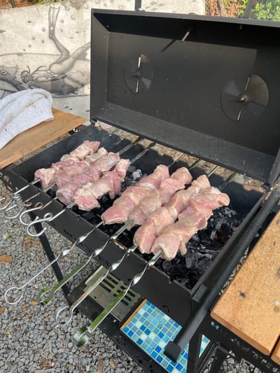The image shows skewers of raw, seasoned meat grilling over hot coals in an outdoor charcoal grill. The grill is black metal and open, with the lid partially raised. Several skewers of meat are arranged on the grill. In the background, a light-colored concrete wall is visible with a faint drawing of what appears to be a frog or other amphibian. A white towel is folded on a wooden surface to the left of the grill. The ground surrounding the grill is covered in small gray gravel. The scene suggests an outdoor cooking activity, possibly in a backyard or recreational area in Sovetskaya Gavan', Russia.