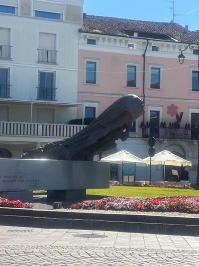 The image shows a sculpture in a city square. The sculpture appears to be a stylized head and neck, made of dark metal, rising from a stone base. The base has text inscribed on it, which appears to be in Italian. In the background, there are buildings with light pink and white facades, some with balconies and windows. There are also two white umbrellas providing shade over outdoor seating areas, suggesting a cafe or restaurant. Flower beds with pink and red flowers are visible in the foreground and around the base of the sculpture. The ground is paved with cobblestones. The scene is illuminated by bright sunlight, casting shadows.

This sculpture is located in Desenzano del Garda, Italy, specifically in Piazza Garibaldi. The inscription on the base reads "I AVIATORI DEL REPARTO ALTA VELOCITA" which translates to "The Aviators of the High Speed Department." The dates "1922 - 1936" are also inscribed, commemorating the existence of this department.