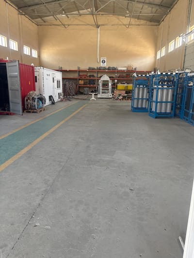 The image depicts the interior of a large warehouse or storage facility. The floor is concrete with visible wear and tear, including cracks and debris. There are several large industrial containers or modular units present, including a red one on the left with its door open, revealing some equipment inside, and a white one next to it with a door and windows.

The back wall of the warehouse is painted a light tan color and shows signs of age. High up on the wall, there are several rectangular windows letting in natural light. The ceiling is a metal truss structure.

Along the back wall, there are multiple metal shelving units stacked with various items, including coiled cables, boxes, and other industrial supplies. In the middle of the warehouse, a white, cage-like structure on wheels is visible. To the right, several tall blue metal racks hold rows of large, silver gas cylinders.

The scene appears to be a storage area for industrial equipment and supplies, possibly related to oil and gas operations, given the context of Baku, Azerbaijan. There are no people visible in the image. The overall impression is one of functional, industrial storage, rather than a display area.