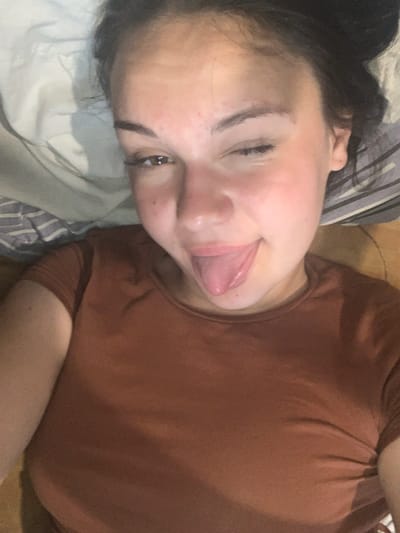 A close-up, overhead, selfie-style shot shows a young woman lying down with her head tilted. Her right eye is open and looking at the camera, while her left eye is squinted. Her tongue is extended out of her mouth. She is wearing a plain, rust-colored t-shirt. The background appears to be a bed with white and grey striped pillows. The lighting is dim, suggesting an indoor setting. The image was taken in Hoholeve, Ukraine.