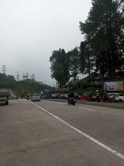 The image shows a congested road with cars, motorcycles, and a bus. A traffic officer stands near parked vehicles. A billboard wishes viewers a "Selamat Hari Raya Idul Fitri". The background includes trees and a cloudy sky. Power lines are visible in the distance. The location is most likely not in Mae Chan District, Thailand, as the signage is in Indonesian.