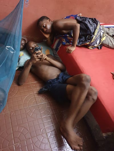 Three individuals are seen relaxing indoors in Idah, Nigeria, captured from an overhead perspective.
In the upper left, one person lies prone on a red mattress or couch, their head turned towards the camera with a neutral expression. They wear light-colored trousers, and their upper body is partially covered by a dark, patterned cloth featuring yellow and blue designs.
In the center and lower right, two other individuals are resting on a light brown tiled floor, which features repeating square and circular patterns. One person is lying on their back, holding a smartphone with both hands. The phone case has faint graphics and possibly text that appears to be "OMG." This individual is shirtless, wearing dark shorts, with their legs bent and bare feet visible on the tiles. Their head is turned towards the camera. Next to them, partially obscured by a blue mosquito net, the head of a third individual is visible, also lying on the floor.
Another pair of bare legs and feet are visible in the upper right quadrant of the image, stretched out on the tiled floor, possibly belonging to a fourth person or an extension of the person on the red mattress. The blue mosquito net indicates a resting or sleeping area. The overall scene suggests a casual and relaxed atmosphere, likely during leisure time.