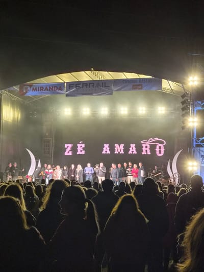 The image shows a stage at an event with a large group of people on it, in Barcelos, Portugal. A vertical sign with the name "Zé Amaro" and a hat logo is displayed prominently. Banners with commercial advertising like "MIRANDA Supermercados" and "FERRAIL" are visible on the left side of the stage. Bright stage lights illuminate the scene and the audience is silhouetted in the foreground. A wagon wheel and crescent moon decorations are above the people on stage. There is a building facade visible behind the stage.