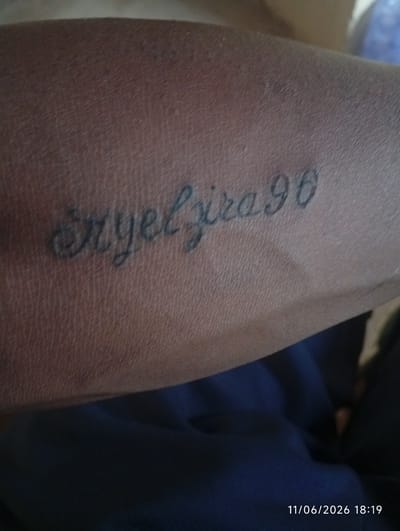 A close-up, slightly blurry shot shows a tattoo on a person's arm. The tattoo, done in black ink, consists of cursive lettering that appears to read "Axelyzira96". The arm is tanned and textured, with visible pores and fine hairs. The background is out of focus, with a dark blue fabric filling the lower portion of the frame. The date and time stamp "11/06/2026 18:19" is visible in the bottom right corner. The location is not specified in the image.