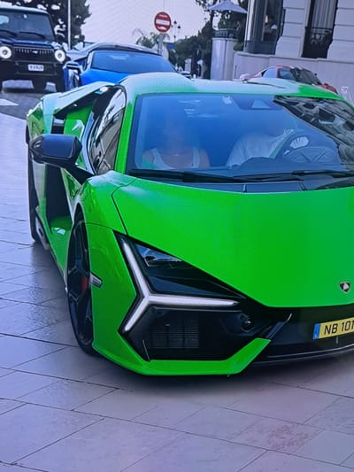 The image shows a vibrant green Lamborghini parked on a tiled street in Cēsis, Latvia. The car is the primary subject, with its sleek design and bright color dominating the frame. The front of the car is visible, featuring sharp, angular headlights and a prominent grille. A yellow license plate with the text "NB 10T" and a blue European Union symbol is attached to the front.
Behind the Lamborghini, other vehicles are partially visible, including a blue car and several black vehicles in the background, suggesting a street or parking area. The setting appears to be urban, with buildings visible in the far background, though they are out of focus. The ground is paved with light-colored tiles, and the lighting suggests it is daytime, possibly with overcast skies as there are no strong shadows.
The overall impression is of a parked luxury car in a European city. There is no clear activity or event taking place, as the car is stationary. The image is a close-up, focusing on the car's design and color, with the surrounding environment providing context.