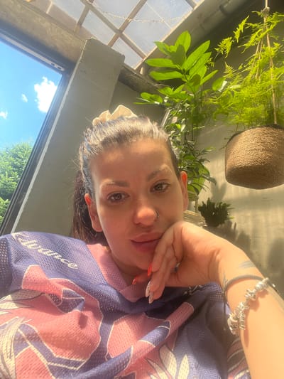This is a close-up, high-angle, eye-level selfie of a young woman in Zelenikovo, Bulgaria. The woman is resting her chin on her hand, looking directly at the camera. She has brown eyes, a nose ring, and her hair is dark brown and pulled back. She is wearing a blue and pink mesh jersey with a floral pattern. She has a bracelet on her left wrist adorned with charms, and her fingernails are painted with orange polish.
The setting appears to be an indoor area with large windows, possibly a greenhouse or a sunroom, given the abundance of lush green plants visible in the background and outside the window. The sunlight streaming in suggests it is daytime, likely morning or afternoon, with bright, clear weather indicated by the blue sky and clouds visible through the windows. The overall mood is relaxed and casual. There is no visible text in the image.