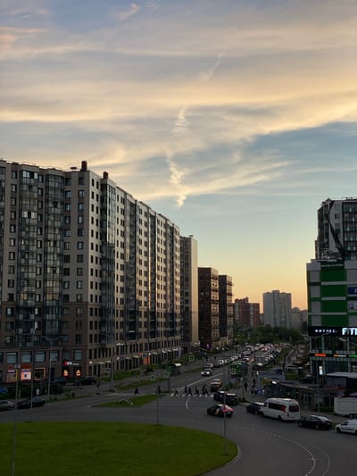 The image shows a street scene in Kudrovo, Russia, during what appears to be either sunrise or sunset, with warm, soft light illuminating the sky and casting long shadows. In the foreground, a large, modern residential building complex dominates the left side of the frame. The complex features multiple stories with a facade of beige and brown panels, interspersed with numerous large windows and balconies. A strip of bright green grass slopes upwards on the far left, suggesting a park or undeveloped area adjacent to the urban development.

To the right of the residential buildings, a multi-lane road runs horizontally across the image, filled with various vehicles, including cars, vans, and what appears to be a bus. Pedestrians are visible on sidewalks and crossing the street at marked crosswalks. Along the right side of the road, commercial buildings are situated. One prominent building is a multi-story structure with a green and white facade, bearing signage that includes "ФИТНЕС" and "FIT". Other commercial establishments with illuminated signs are visible, indicating shops and services. Further down the road, more residential buildings of varying heights and architectural styles are visible in the distance, fading into the hazy sky. The sky itself is a pale blue, transitioning to shades of yellow and orange near the horizon, with wispy clouds. The overall scene depicts a bustling urban environment with a mix of residential and commercial spaces, characterized by modern architecture and active street life under a diffused sky.