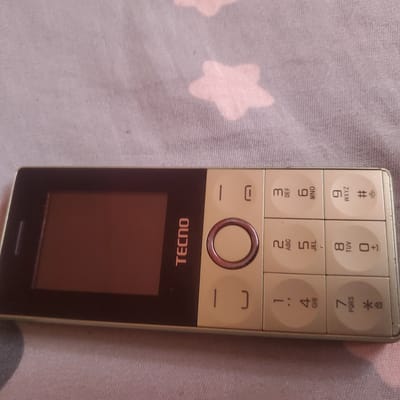 The image shows a close-up of a Tecno feature phone, lying on a pink fabric with a subtle star pattern. The phone is predominantly light beige with a dark brown screen on the left side. The keypad is visible, with numbers and letters arranged in the standard phone layout. Notably, the brand name "TECNO" is printed vertically in orange on the dark screen area. The keys include numbers 1 through 9, as well as a star and hash key, with corresponding letters or symbols printed below the numbers. A circular navigation button is centered above the keypad. The phone appears to be in good condition. The setting suggests an indoor environment, likely a bed or sofa, given the fabric. There are no people visible in the image. The lighting appears to be soft, possibly from an indoor light source, and there are no indications of the time of day or specific weather conditions. The location context of Ribina East, Nigeria, is provided but cannot be directly inferred from the visual elements of the image.