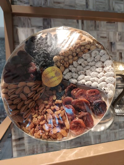 A circular platter of assorted dried fruits and nuts is displayed on a glass shelf. The platter is divided into sections, each containing a different item. Visible contents include almonds, raisins, dried apricots, pistachios, dark dried fruits that appear to be prunes or dates, and small white confectionery items that resemble sugared nuts. A round label with an illustration of buildings and the text "UZBEKISTAN SAMARKAND" is adhered to the center of the platter. The platter is covered in clear plastic packaging. The background consists of a wall with a patterned tiled surface and a wooden shelf. The lighting is bright, creating reflections on the plastic packaging and the glass shelf. The overall scene suggests a marketplace or a shop in Konigil, Uzbekistan, where such food items are sold.