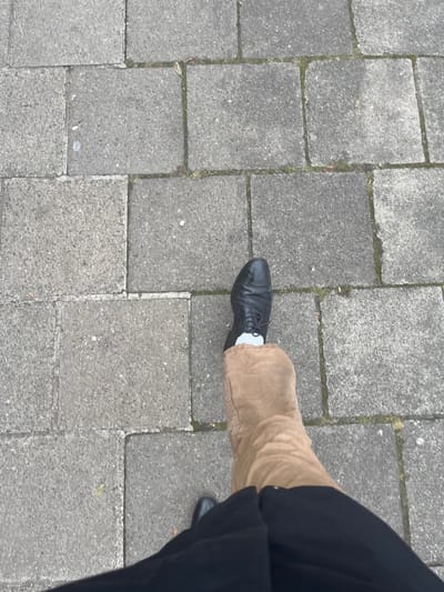 The image, captured from an overhead perspective, focuses on the lower legs and feet of a person standing on a paved outdoor surface. The pavement is composed of rectangular, grey concrete or stone blocks laid in a grid pattern. Thin lines of dirt and small patches of moss are visible in the crevices between the blocks.
The left foot of the person is prominently featured, wearing a black, lace-up leather shoe. A segment of a white sock is visible between the top of the shoe and the hem of a pair of tan-colored trousers, which appear to be made of a sturdy, possibly corduroy or canvas, fabric. The trousers show natural folds and creases. The right foot, also in a black shoe, is partially visible towards the bottom right corner of the frame, largely obscured by a dark, possibly black, garment that covers the leg, likely another pair of trousers or an overcoat.
The lighting is soft and even, suggesting an overcast day. No harsh shadows are present. Scattered across the paving stones are a few small pieces of debris. The scene depicts a common urban setting, likely a sidewalk or pedestrian area in a city such as Bamberg, Germany. There is no visible text in the image.
