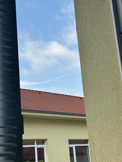 The image captures an upward-looking view, rotated 90 degrees clockwise, creating an abstract perspective of an urban setting under a clear sky.

Framing the scene, a light beige or pale yellow wall with a coarse, stucco-like texture runs horizontally across the top of the image. A dark, possibly black or charcoal grey, slatted or ribbed structure forms a horizontal band across the bottom.

Between these framing elements, a bright blue sky with scattered white, wispy clouds fills the left and central parts of the frame. To the right, a building is visible. It features a distinct red tiled roof with a wavy pattern, sloping downwards towards the right. A dark gutter pipe runs along the edge of this roof. Below the roofline, a segment of the building's facade, painted a similar light yellow color to the textured wall at the top, is visible. Two windows with white frames are inset into this facade, reflecting internal darkness or shadows.

The scene depicts a daytime setting with fair weather. No people or specific activities are visible, and there is no discernible text. The architecture, particularly the red tiled roof and stucco walls, suggests a location in Southern or Eastern Europe, consistent with the provided context of Elena, Bulgaria.
