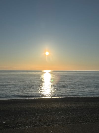 The image shows a sunrise over the Black Sea in Kobuleti, Georgia. The sun is visible in the sky, and its reflection can be seen on the surface of the water. The shoreline is visible on the right side of the image. The sky is a light blue color, and the water is a darker shade of blue. The image appears to be taken at ground level.