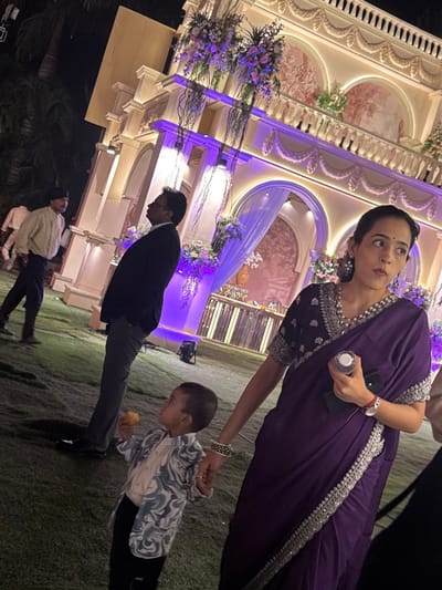 A woman in a purple sari holds a water bottle and the hand of a small child in a patterned shirt. The child is eating something. Behind them is a man in a suit. In the background is a large, ornately decorated building, possibly for a wedding, lit with purple lights. A person in a light-colored outfit is partially visible in the lower left corner. They are standing on grass.