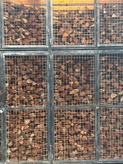 The image shows a close-up of nine wire mesh containers filled with brown, dried wood pieces. The containers are arranged in a 3x3 grid and appear to be part of a larger storage system. The frames of the containers are dark gray, and the wire mesh is a light tan color. The wood inside is stacked densely within each container. The background is indistinct. Based on the metadata, this is located in Câmara de Lobos, Portugal.